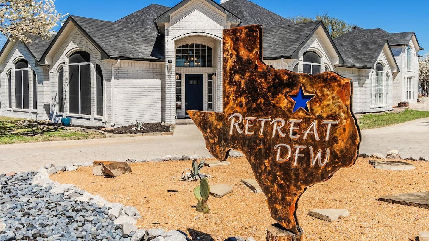Front elevation of the main house with a Texas-shaped 'Retreat DFW' sign in the landscaping